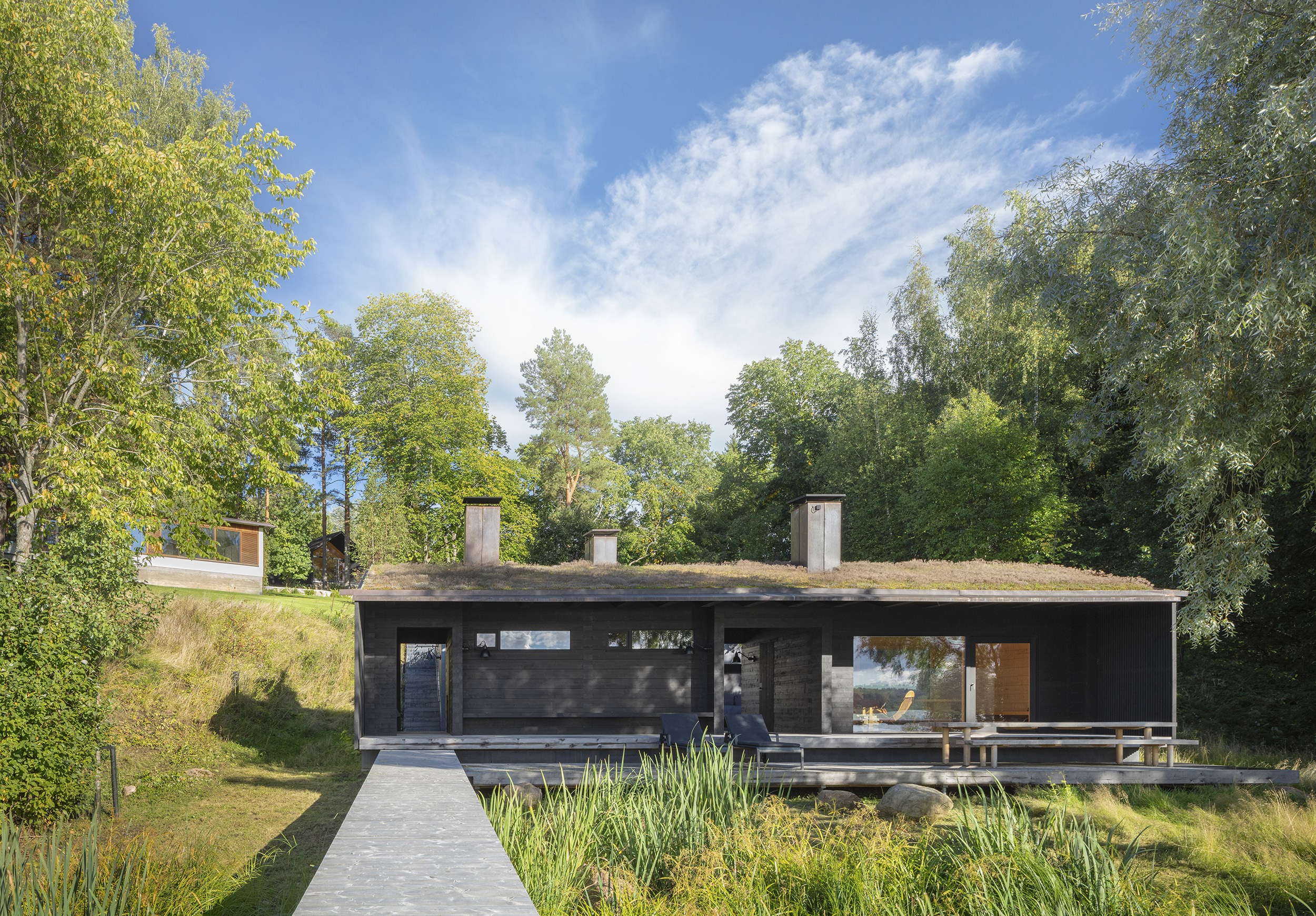 A wooden sauna by the lake amidst a green landscape, with the main building up on the hill in the background and a wooden boardwalk in the foreground