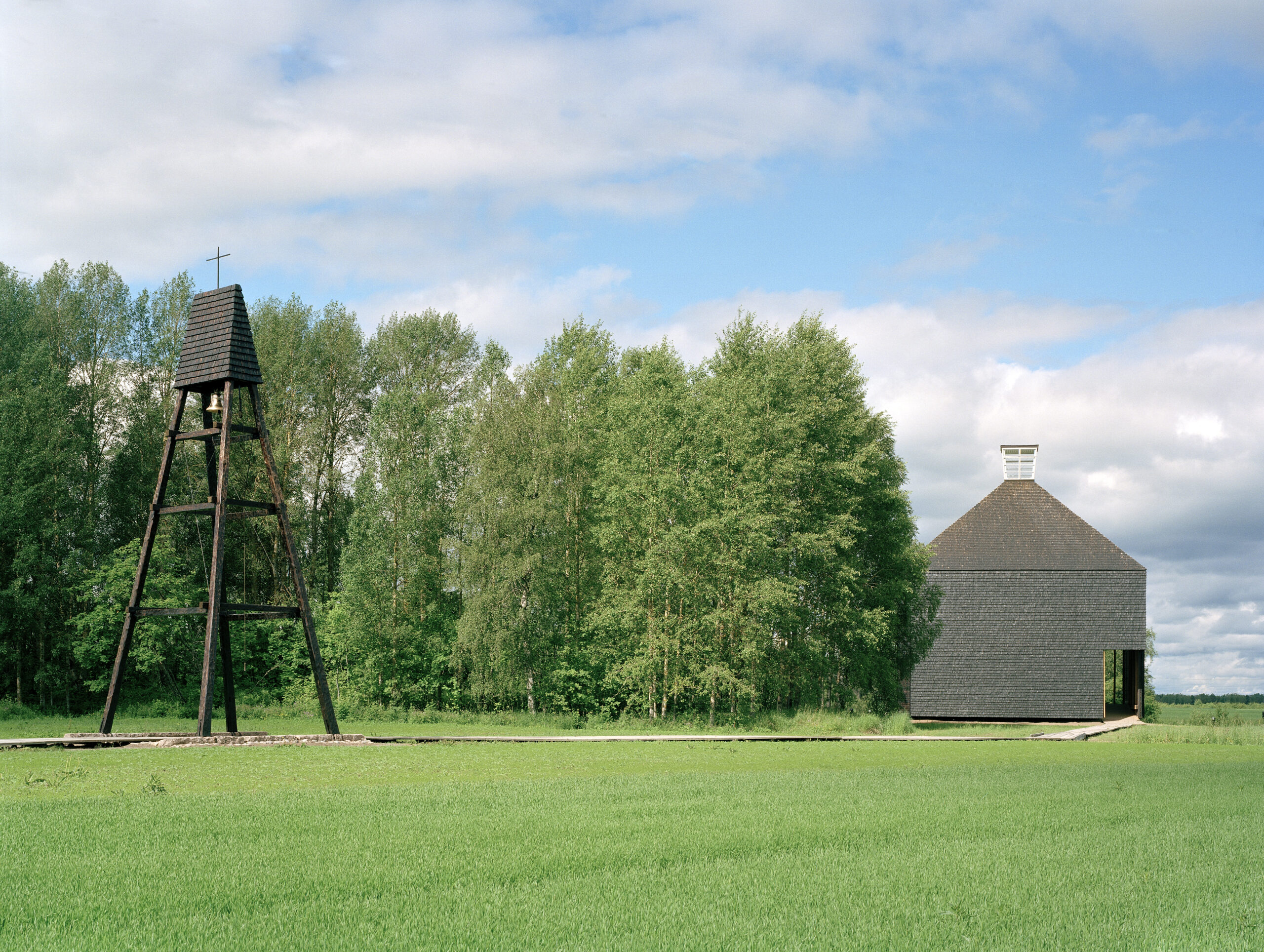 A view showing a bell tower and a church clad with shingles sitting on the edge of a forest