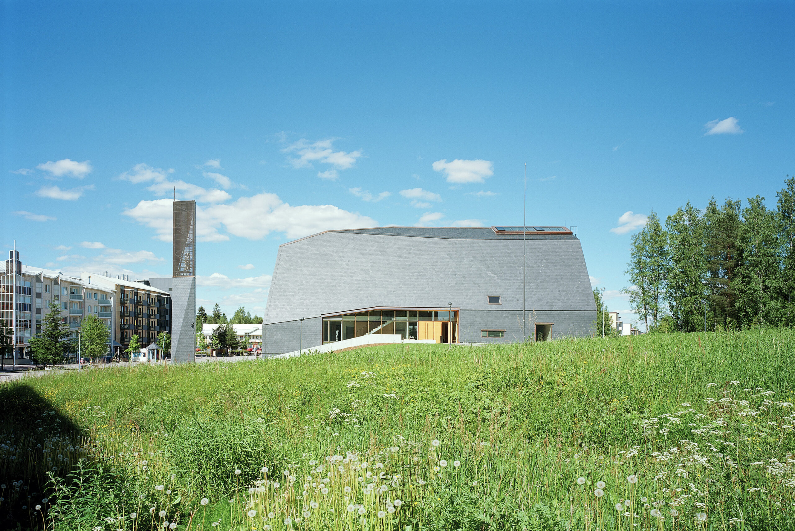 View of a church and a bell tower clad in slate stone with a field of grass in the foreground and a block of housing in the background